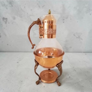 May include: A glass carafe with a copper-coloured metal base, lid, and handle. The metal components feature an ornate, filigree-style band around the neck and decorative, curved legs. The carafe has a clear glass body and a small, pointed finial.