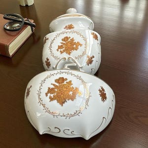 Puede incluir: Dos tarros de cerámica blanca con tapas, adornados con diseños florales dorados y ribetes. Los tarros están apilados, mostrando una estética decorativa y elegante. Las tapas tienen pequeños pomos para facilitar el manejo.