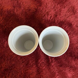 Pu&ograve; includere: Due contenitori in ceramica bianca di forma cilindrica con un'ampia apertura. I contenitori sono appoggiati su uno sfondo rosso strutturato. L'interno dei contenitori &egrave; grigio chiaro.