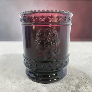May include: A dark red glass tumbler with a detailed profile of a person embossed on the side. The top and bottom rims feature star and dot patterns. The tumbler is a cylindrical shape.