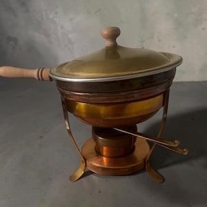 May include: A copper fondue pot with a brass lid and a wooden handle. The pot sits on a metal stand with a brass band and a central burner. The lid features a small wooden knob handle. The entire assembly is constructed from metallic components.