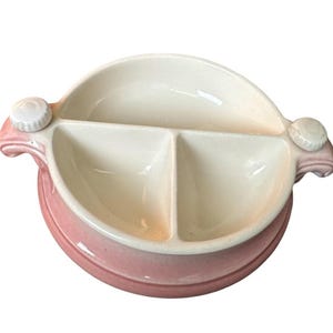 Puede incluir: Un plato de cerámica dividido con una base rosa y un interior blanco. El plato tiene dos asas con un botón decorativo a cada lado. El interior está dividido en tres secciones, perfecto para servir aperitivos o porciones pequeñas.