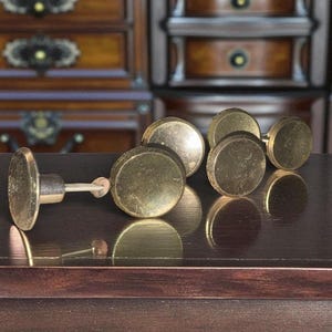 May include: A collection of antique brass cabinet knobs with a round, flat design. The knobs are arranged on a dark brown surface, reflecting the warm, golden tones of the metal. The background features a wooden cabinet.