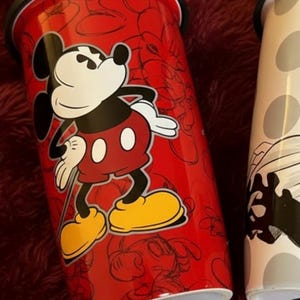 Pu&ograve; includere: Bicchiere da viaggio rosso con Topolino. Il bicchiere ha un coperchio nero e un design di Topolino bianco, nero e rosso. Lo sfondo del bicchiere presenta un motivo a contorno di Topolino. &Egrave; visibile anche un secondo bicchiere con un design in bianco e nero.