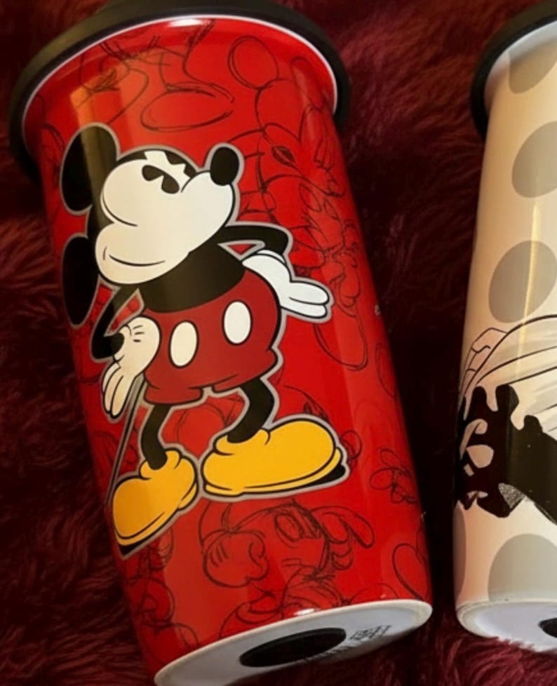 Pu&ograve; includere: Bicchiere da viaggio rosso con Topolino. Il bicchiere ha un coperchio nero e un design di Topolino bianco, nero e rosso. Lo sfondo del bicchiere presenta un motivo a contorno di Topolino. &Egrave; visibile anche un secondo bicchiere con un design in bianco e nero.