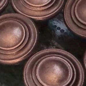 Könnte beinhalten: Nahaufnahme mehrerer kupferfarbener, kreisförmiger Schrankknöpfe. Jeder Knopf hat ein erhabenes, konzentrisches Design, das einen geschichteten Effekt erzeugt. Die Knöpfe haben einen warmen, metallischen Glanz, der auf einen Vintage- oder Antik-Look hindeutet. Der Hintergrund ist dunkel und gefleckt.