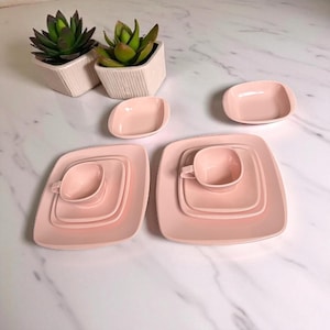 May include: A collection of pink ceramic dishware, including square plates, small bowls, and teacups. The set is arranged on a white surface, with two small potted succulents in the background. The dishware has a retro, mid-century modern aesthetic.