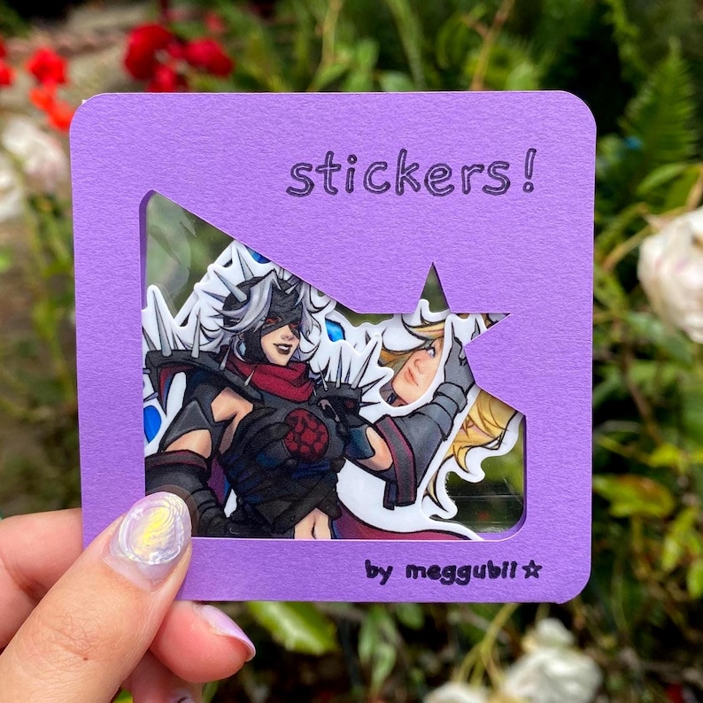 May include: A purple card with the word "stickers!" and a star-shaped cutout, displaying two character stickers. The stickers feature detailed illustrations with a dark color palette. The text "by meggubii" is at the bottom.