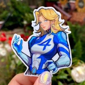 May include: A sticker featuring a female superhero with blonde hair and blue eyes. She wears a blue and white uniform with a large number 4 on the chest. The sticker has a white border and is held by a hand with a colorful manicure.