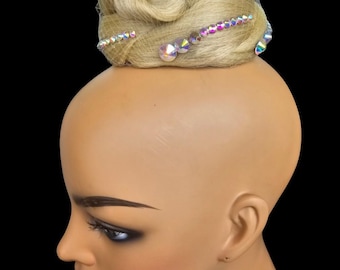 May include: A blonde hairpiece with sparkling iridescent rhinestones is displayed on a mannequin head. The hair is styled in a bun with the rhinestones arranged in a decorative pattern. The mannequin has a neutral skin tone and is set against a black background.