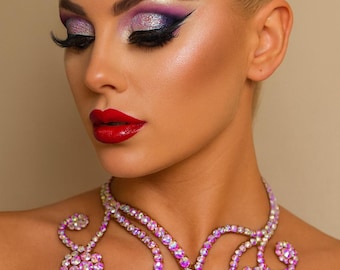 May include: A woman with blonde hair and dramatic eye makeup, wearing a sparkling pink and purple rhinestone necklace. The necklace has a floral design and is set against a neutral background.