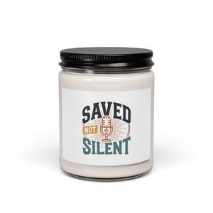 Puede incluir: Un tarro de vela blanco con tapa negra. La etiqueta presenta las palabras "SAVED NOT SILENT" en un diseño audaz y multicolor, con un gráfico de micrófono y una cruz.