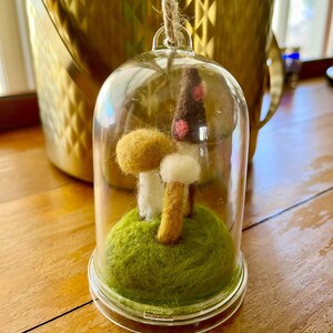 May include: A clear glass cloche with a small felt mushroom scene inside. The scene includes three felt mushrooms on a green felt base. The cloche is suspended by a twine loop. The background includes a gold-colored bucket.