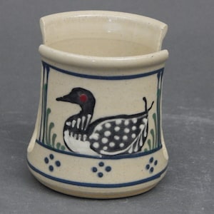 May include: A ceramic wall pocket with a beige speckled finish. The pocket features a blue rim and a painted design of a loon bird with black, white, and red details. Green and blue accents surround the bird, and blue dots decorate the base.