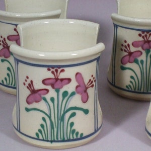 May include: Set of four white ceramic utensil holders with a floral design. Each holder features pink flowers with green stems and leaves on a white background with a blue border.