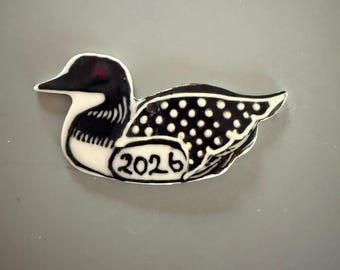 May include: A black and white ceramic loon ornament featuring a red eye, a white polka dot pattern on the wing, and the year 2026 printed in black ink on the lower body section. The bird is depicted in a side profile view with a white neck and chest.