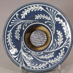 May include: A blue ceramic flower frog with a white leaf design around the rim. The center has a brass-colored ring with a metal pin holder for arranging flowers. The frog is approximately 6 inches in diameter.