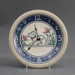 May include: A white ceramic clock with a blue rim and a dog design. The clock face has black Roman numerals and black hands. The dog is white with a pink flower and green leaves.