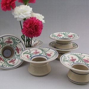 May include: Set of four ceramic flower frog vases with a white background and floral designs in pink, green, and blue. The vases are designed to hold flowers in a traditional Japanese ikebana style.