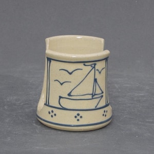 May include: A cream-colored ceramic napkin holder featuring a hand-painted blue sailboat and bird design. The cylindrical piece has a wide opening at the top, a flared base with blue dot accents, and a cutout on the side for easy access to napkins.