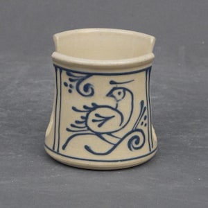 May include: A cream-colored ceramic sponge holder with a cylindrical shape and a notched top rim. The exterior features a hand-painted dark blue line drawing of a bird perched on a branch, accented with decorative swirls and dots on the sides.