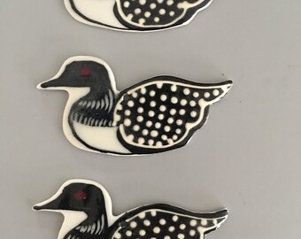 May include: Three decorative loon figurines. Each loon has a black head and beak, a white body, and black and white dotted wings. The loons have a red eye and are arranged vertically against a gray background.