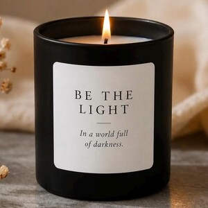 May include: A matte black cylindrical candle in a glass container with a lit wick. A white rectangular label on the front features the black text: BE THE LIGHT. In a world full of darkness. The candle is positioned upright with a visible flame.