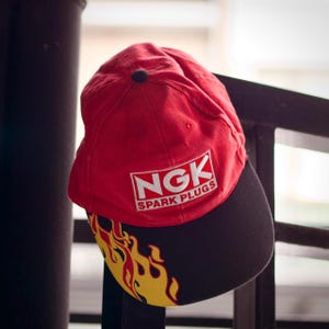 Może przedstawiać: Czerwono-czarna czapka z daszkiem z wzorem płomieni na daszku. Na czapce znajduje się białe logo z napisem "NGK SPARK PLUGS".