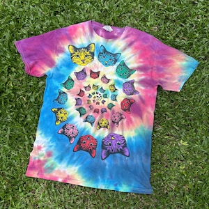 Könnte beinhalten: Ein Batik-T-Shirt mit einem Spiralmuster aus Katzenköpfen in verschiedenen Farben. Der Hintergrund des Shirts zeigt eine Mischung aus Lila, Rosa, Blau und Gelb. Die Katzenköpfe sind gelb, blau, grün, lila und rot.