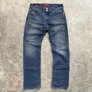 Könnte beinhalten: Blaue Jeans aus Denim mit verwaschener Optik. Die Jeans haben ein klassisches Fünf-Taschen-Design, einen Knopf- und Reißverschluss und ein rotes Logo-Etikett über der rechten Gesäßtasche. Die Jeans liegen flach auf einer Betonoberfläche.