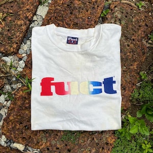 May include: White t-shirt with the word "fuct" in a gradient of red, pink, yellow, and blue. The shirt has a small "Fuct" logo on the collar. The shirt is made of a soft material.
