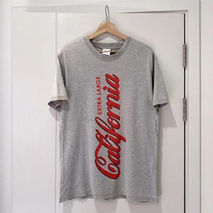 May include: A heather gray short-sleeve t-shirt featuring the red text California written vertically in a script font, with the words EXTRA LARGE printed in a smaller red sans-serif font next to it. The shirt is displayed on a wooden hanger.