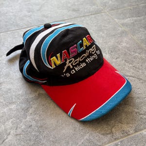 May include: Black NASCAR Racing baseball cap with a red bill and blue and white accents. The cap features embroidered text that reads "NASCAR Racing" and "it's a kids thing to".