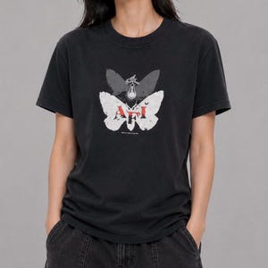 May include: A black short-sleeve t-shirt featuring a graphic of two overlapping butterflies. The top butterfly is dark gray, while the bottom one is white with the red text AFI printed across it. A small lightbulb icon is centered above the text.