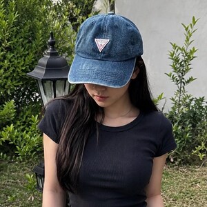 May include: A blue denim baseball cap with a distressed finish and a triangular white patch on the front featuring the red text GUESS. The cap has a curved brim, stitched eyelets on the crown, and a structured front panel with a classic six-panel design.