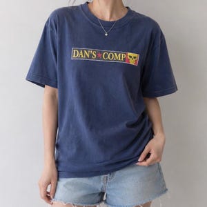 May include: A navy blue short-sleeve t-shirt featuring a rectangular graphic on the chest with the text DAN'S COMP in yellow letters, a small red star, and a yellow skull icon. The shirt has a crew neckline and is paired with light blue denim shorts.