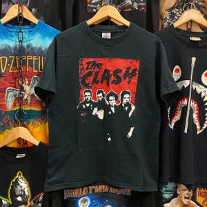 May include: Black t-shirt with a red and white graphic of The Clash band members. The band's name is printed in large white letters on a red background.