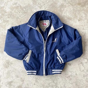 May include: A navy blue vintage jacket with a white collar and cuffs. The jacket has a zipper closure and features the word "TOUCH DOWN" on the inside collar. The jacket also has a logo of a football player.
