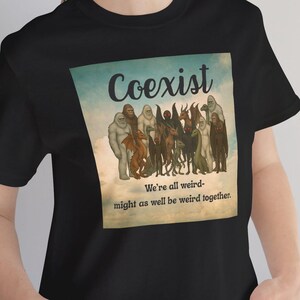 May include: Black t-shirt with a graphic that says "Coexist" above a group of cryptids. The text "We're all weird - might as well be weird together" is below the cryptids. The shirt is made of a soft material.