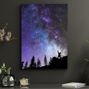 May include: A canvas print featuring a vibrant depiction of the night sky, showcasing a gradient of purple, blue, and white hues. A silhouette of a person with arms raised stands atop a mountain, with dark trees in the foreground.