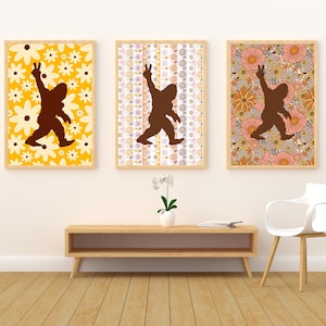 May include: Three framed art prints featuring a brown silhouette of Bigfoot making a peace sign. Each print has a different floral background in yellow, white, and orange tones. The prints are displayed above a wooden console.