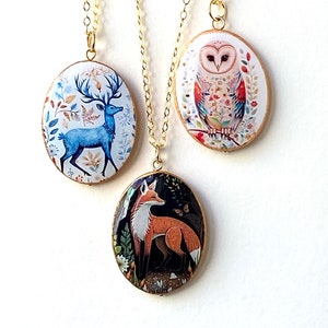 May include: Three gold-tone necklaces with oval pendants featuring woodland animal designs. The pendants showcase a blue deer, a colorful owl, and a red fox. Each pendant hangs from a delicate gold chain, creating a charming and nature-inspired accessory.