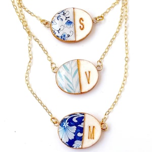 Könnte beinhalten: Drei goldene Kettenhalsbänder mit ovalen Anhängern. Jeder Anhänger ist zur Hälfte weiß mit einem goldenen Initial und zur Hälfte aus blau-weiß gemustertem Porzellan. Die Initialen sind S, V und M.