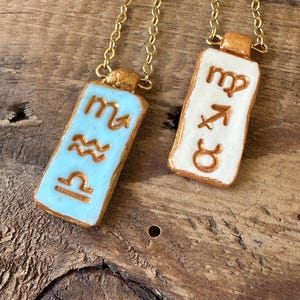 May include: Two gold-colored chain necklaces with rectangular pendants. One pendant is light blue with gold-colored zodiac symbols, and the other is white with gold-colored zodiac symbols. The pendants are rectangular with rounded edges.