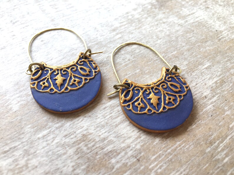 May include: A pair of blue and gold hoop earrings with a delicate, intricate design. The earrings are made of polymer clay and have a gold-toned wire hoop.