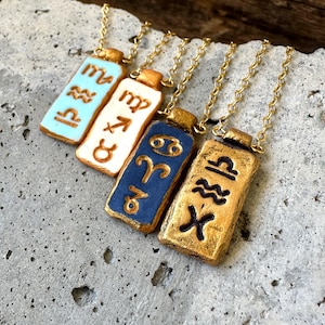 May include: Four rectangular pendant necklaces with gold chains. Each pendant features a different zodiac symbol in gold against a background of light blue, white, dark blue, and gold. The pendants are displayed on a textured gray surface.