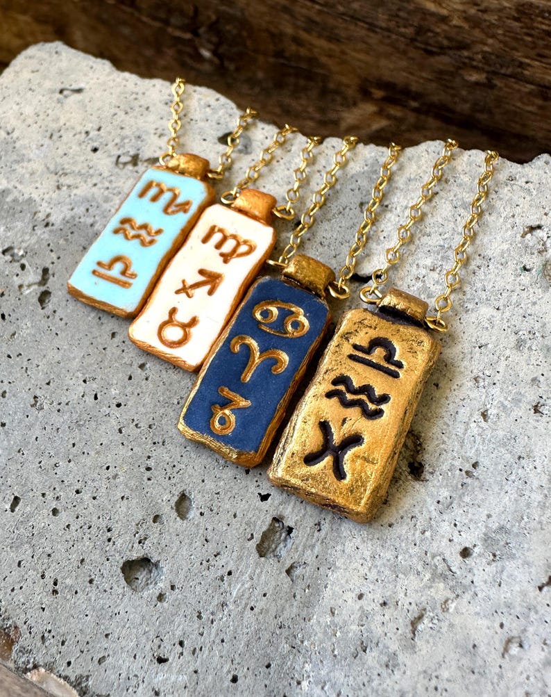 May include: Four rectangular pendant necklaces with gold chains. Each pendant features a different zodiac symbol in gold against a background of light blue, white, dark blue, and gold. The pendants are displayed on a textured gray surface.