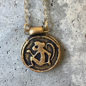 May include: A gold-colored pendant necklace featuring a circular charm with a monkey design. The monkey is depicted in a stylized, seated pose. The pendant hangs from a delicate gold chain, creating a simple yet eye-catching piece of jewelry.