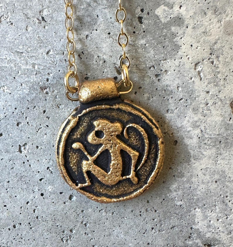May include: A gold-colored pendant necklace featuring a circular charm with a monkey design. The monkey is depicted in a stylized, seated pose. The pendant hangs from a delicate gold chain, creating a simple yet eye-catching piece of jewelry.