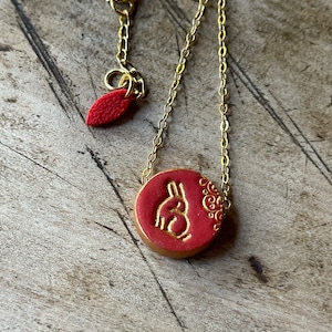 May include: A gold chain necklace with a red circular pendant featuring a gold rabbit silhouette and a gold floral design.